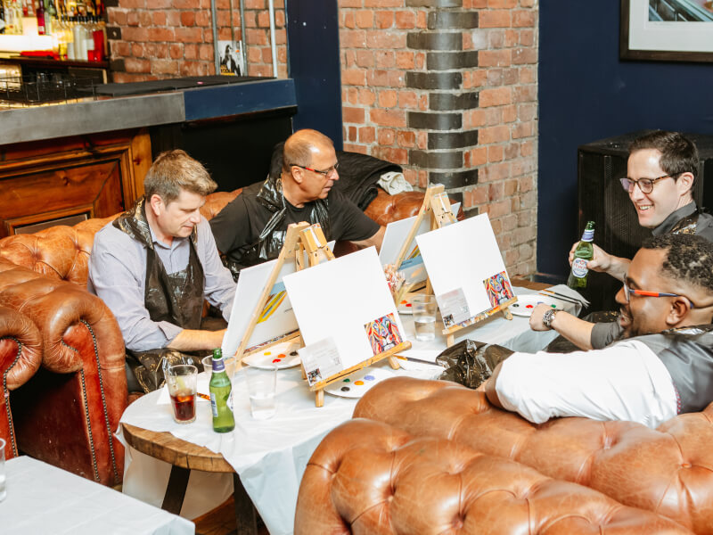Four seated men at a painting class, each with a canvas